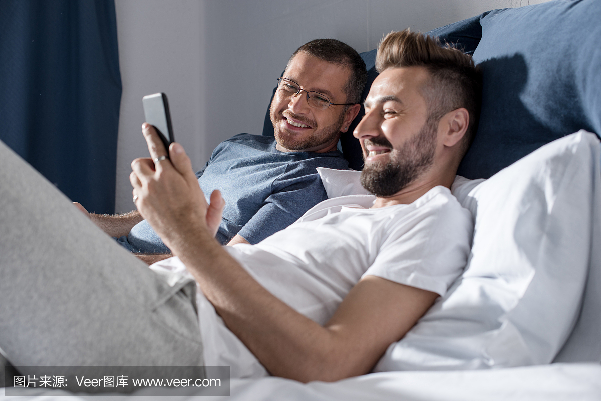 快乐的同性恋夫妇在躺在床上时使用智能手机