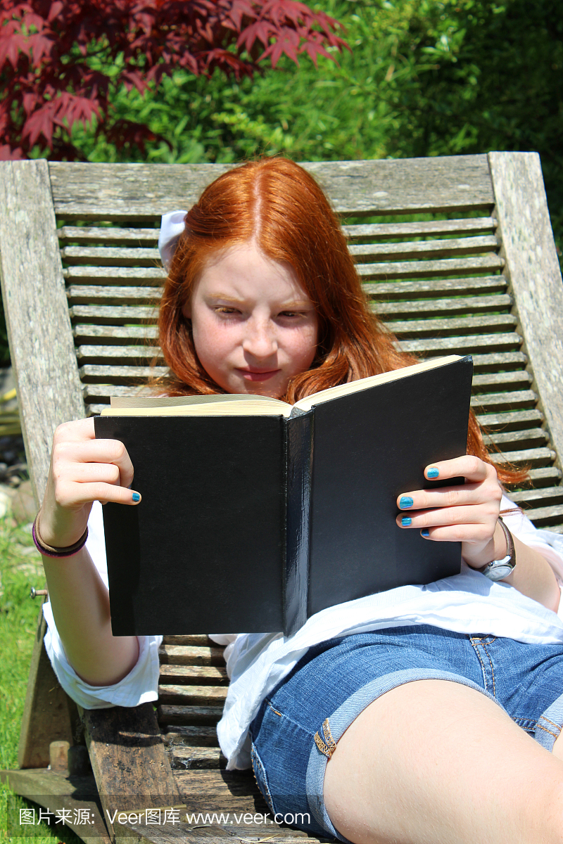 年轻女孩阅读书,木制花园sunlounger \/蒸笼的形