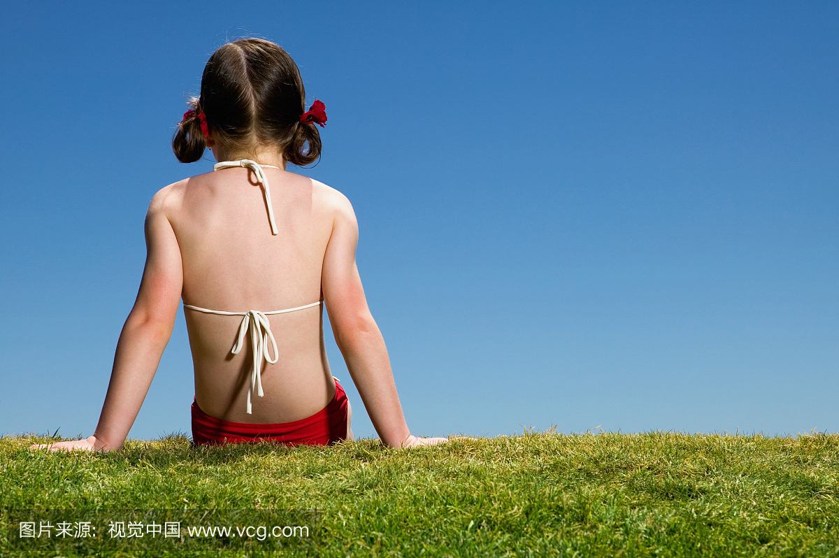 穿着比基尼泳装的女孩(6-8)坐在草地上,后视图