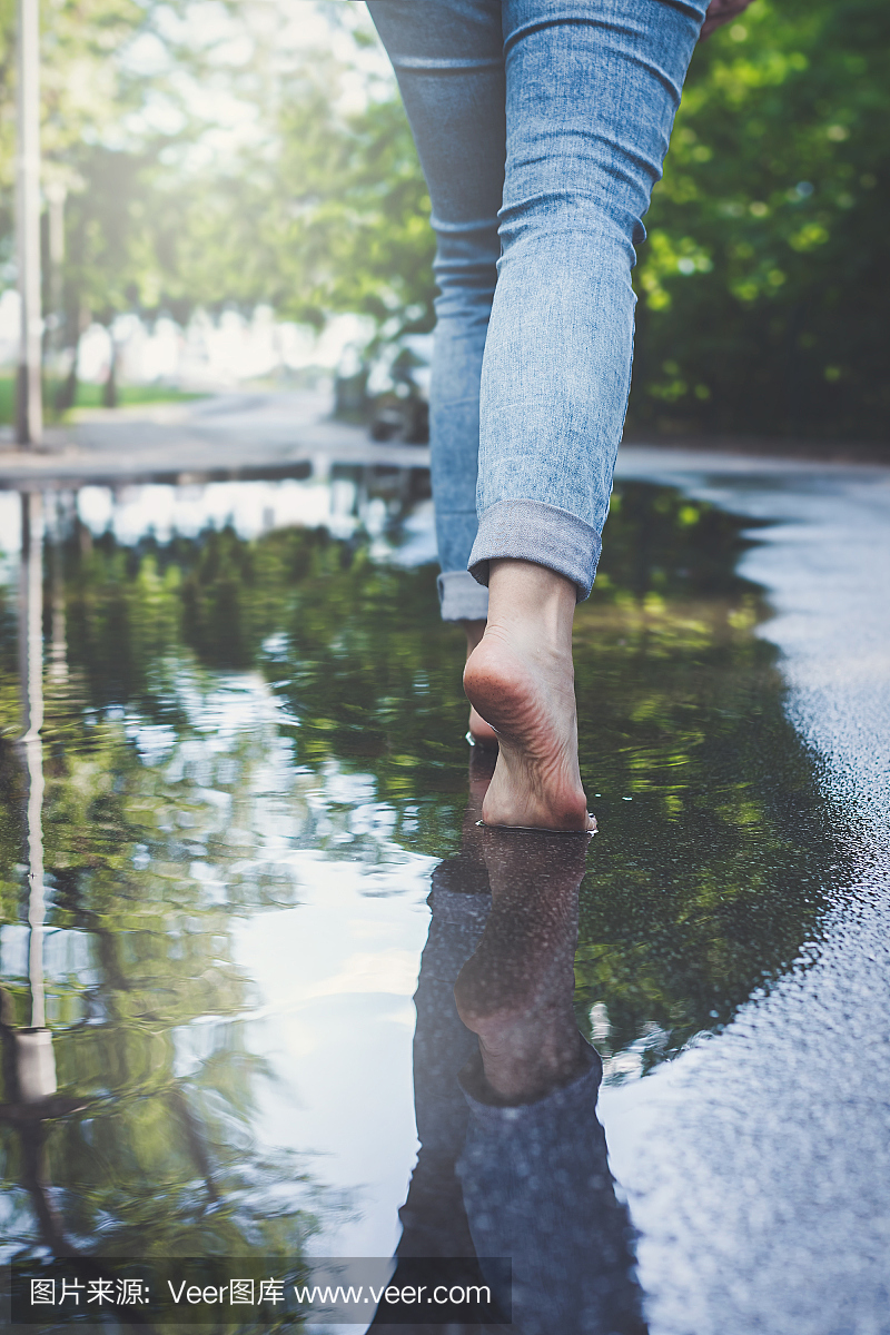 蓝色牛仔裤的赤脚女子在城市环境中穿过水坑