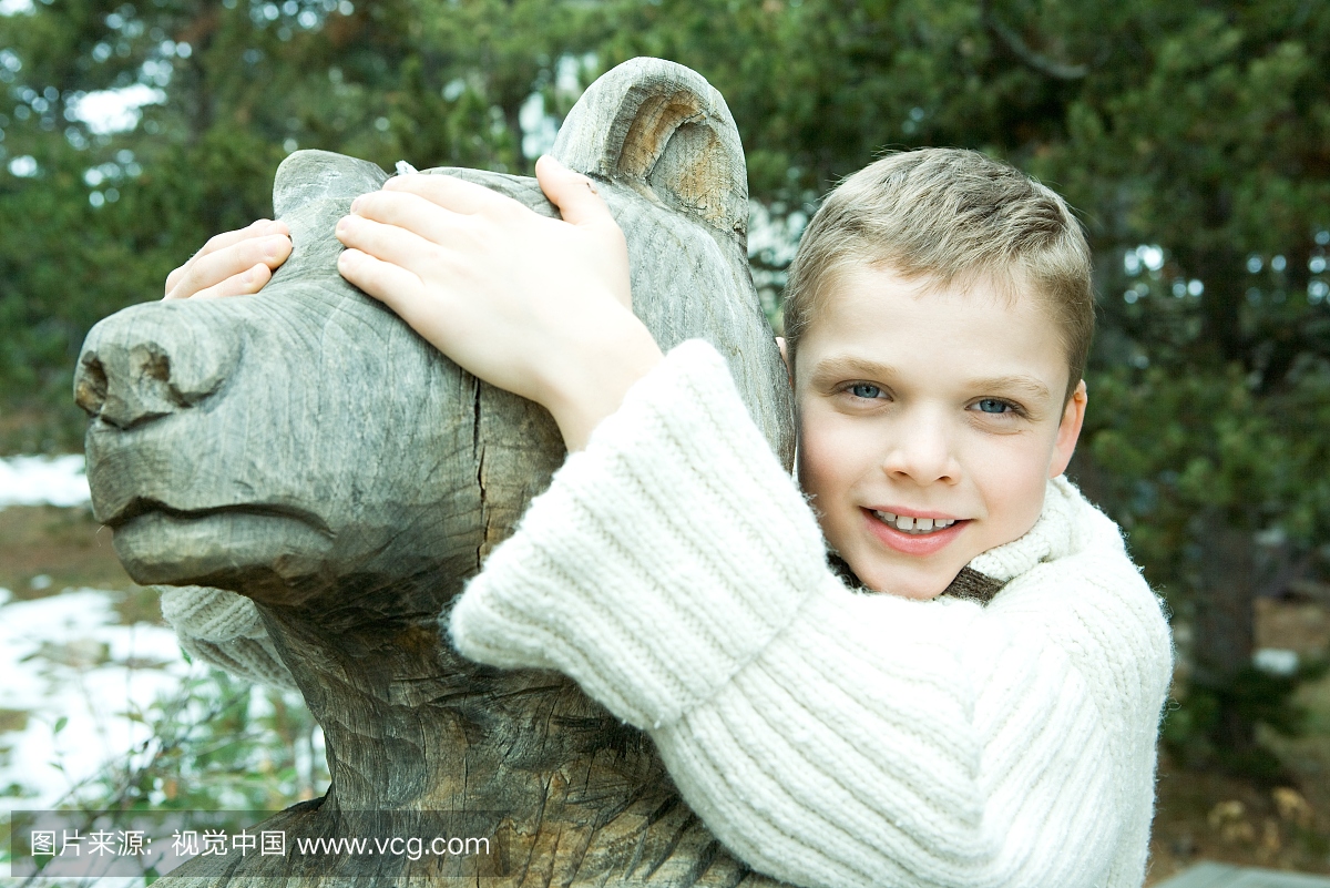 男孩拥抱熊雕像,用手遮住眼睛,微笑着相机
