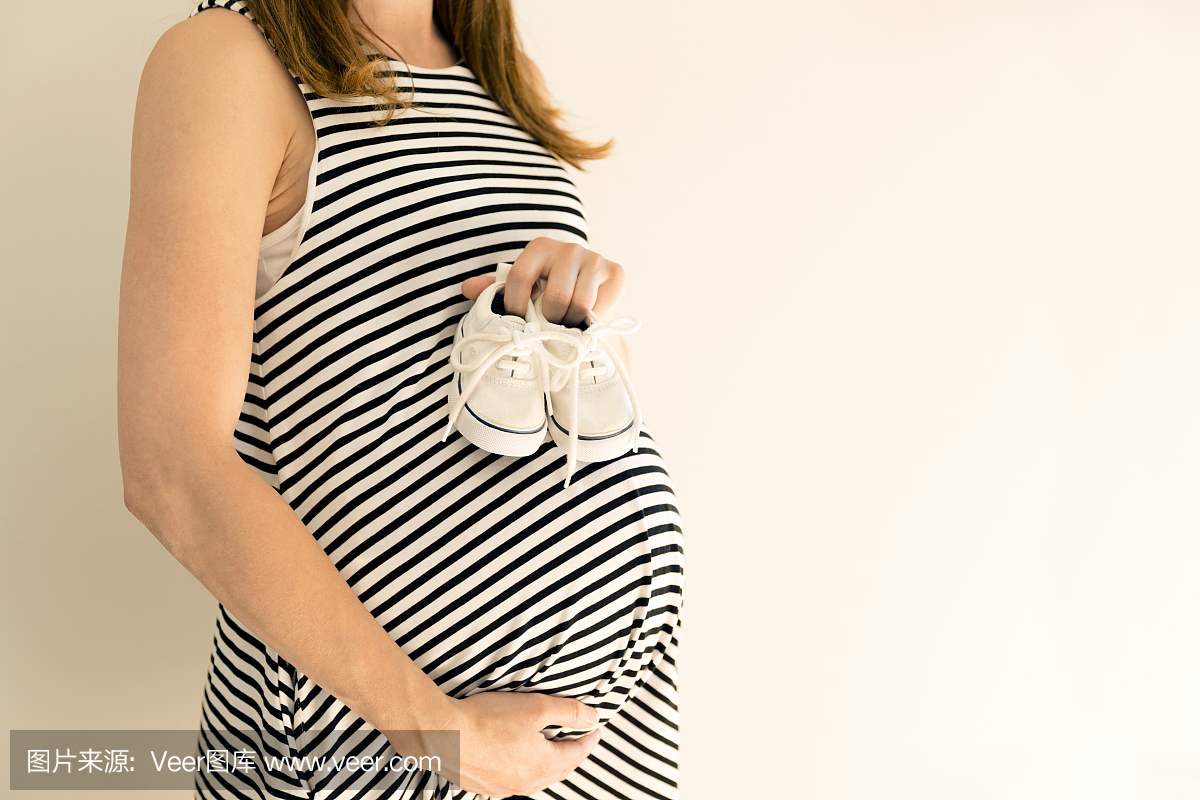 孕妇在她的肚子上抱着婴儿鞋。怀孕和婴儿的概