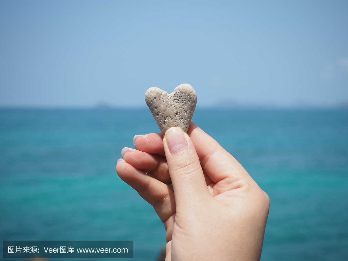 拿着心脏形状的石头在蓝色夏天沙滩背景的女人