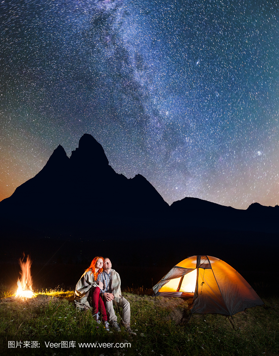 徒步旅行者在晚上坐在篝火附近和发光的帐篷